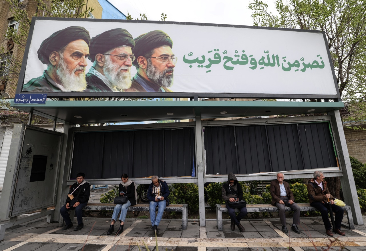 Iranian sit at bus stop beneath a billboard depicting Iran's late supreme leader Ayatollah Ruhollah Khomeini, from left, his successor the late Ayatollah Ali Khamenei and the new supreme leader, Mojtaba Khamenei in Tehran. Iranian sit at bus stop beneath a billboard depicting Iran's late supreme leader Ayatollah Ruhollah Khomeini, from left, his successor the late Ayatollah Ali Khamenei and the new supreme leader, Mojtaba Khamenei in Tehran.